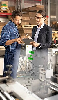 A man and woman discuss a digital workflow on a tablet in a manufacturing environment, with machinery in the background.