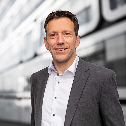 Jürgen Rieger, Executive Manager Consulting, steht in einem grauen Anzug lächelnd vor einem modernen Gebäude.