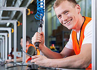 Smiling technician in an orange vest using a power tool to assemble components in a workshop setting.